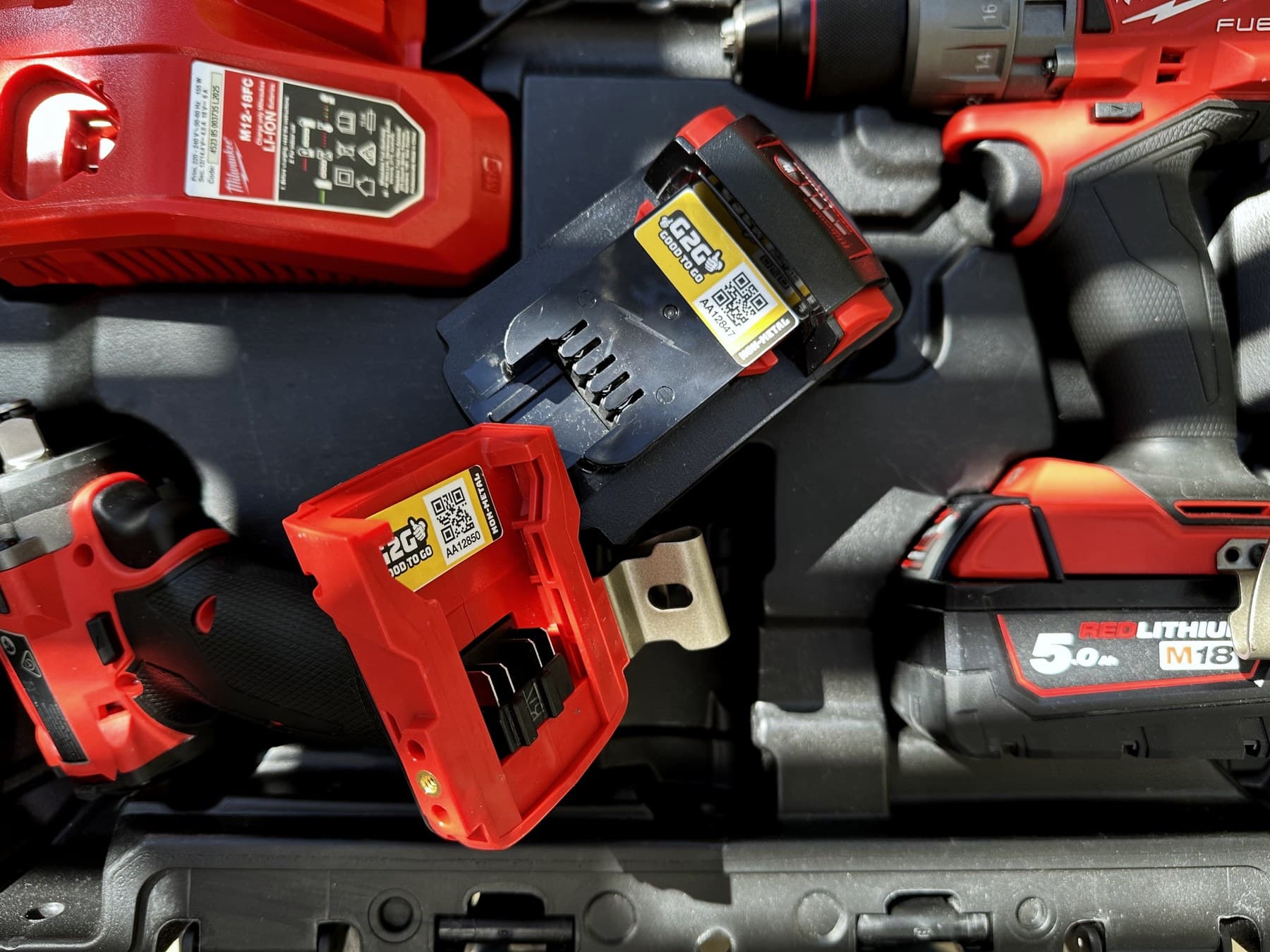 Milwaukee power tools, batteries and a charger in their case. Two of the batteries carry a yellow G2G tag with a QR code on it.